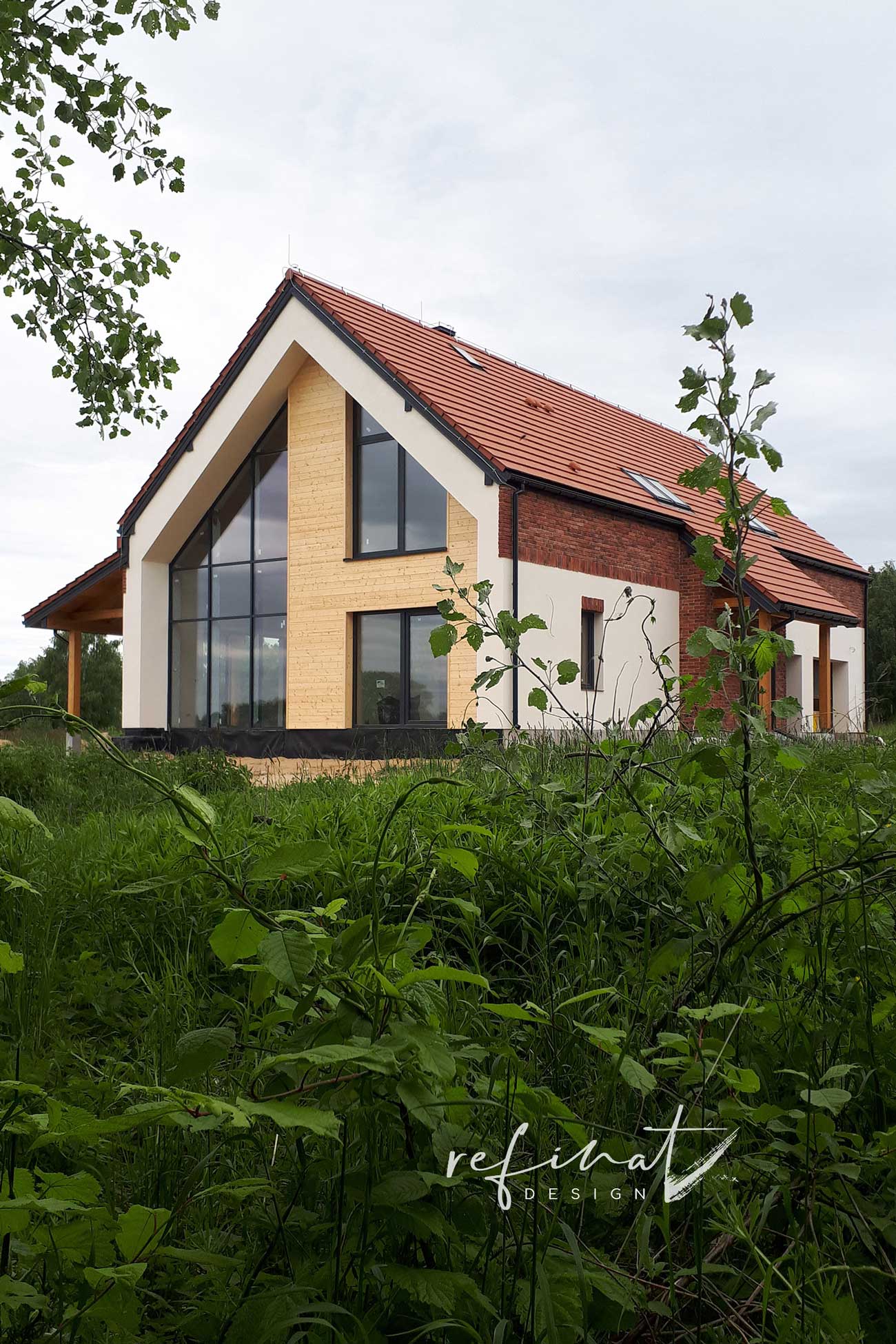 Dom z czerwonym dwuspadowym dachem i dużym szkleniem na froncie, pokryty cegłą i białym tynkiem, w zieleni